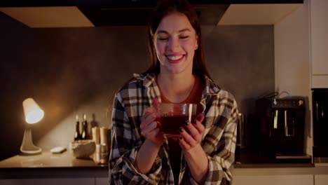retrato de una chica morena feliz y alegre en una camisa a cuadros que sopla en una taza de té caliente en sus manos en un apartamento moderno en la cocina por la noche