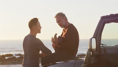 Happy-caucasian-gay-male-couple-sitting-on-car-playing-guitar-and-talking-at-the-beach