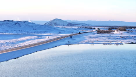 Camino-Invernal-Que-Serpentea-A-Través-Del-Nevado-Paisaje-Volcánico-De-Myvatn-Al-Atardecer,-Con-Un-Lago-Tranquilo-Y-Una-Luz-Suave