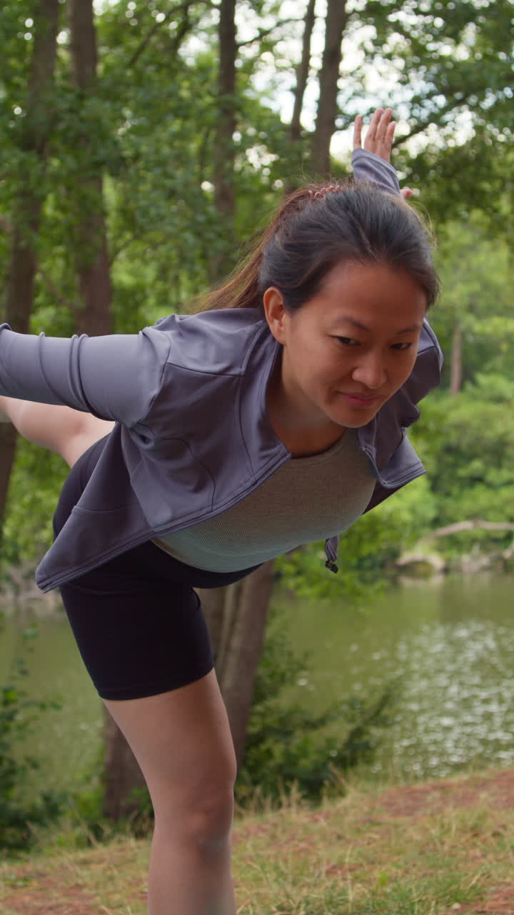 스포츠 옷을 입고 요가를 하는 여성의 수직 비디오, 호수 또는 강 의 숲에서 균형을 잡고 창, 자연의 평화와 아름다움을 즐기고, 실시간으로 촬영