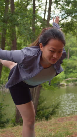 video vertical de una mujer con ropa deportiva haciendo yoga estirándose y equilibrándose en el bosque junto a un lago o río disfrutando de la paz y la belleza de la naturaleza filmado en tiempo real