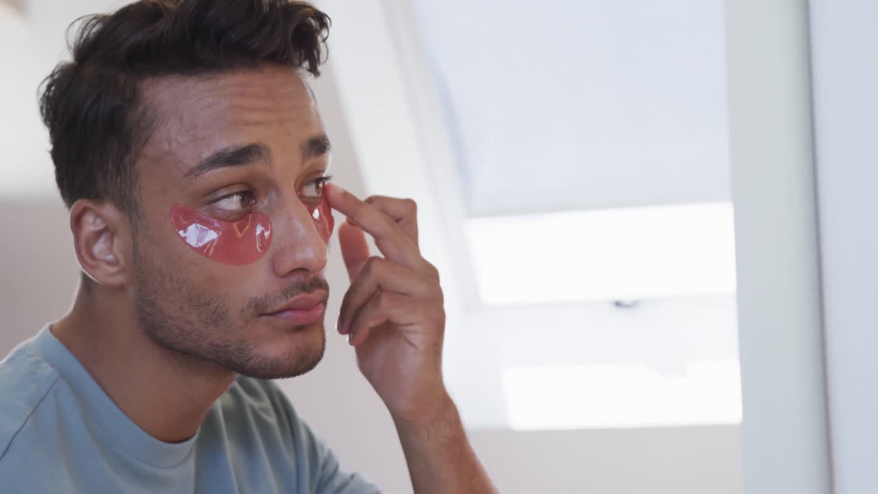 Biracial man looking in mirror applying under eye masks in bathroom, slow motion