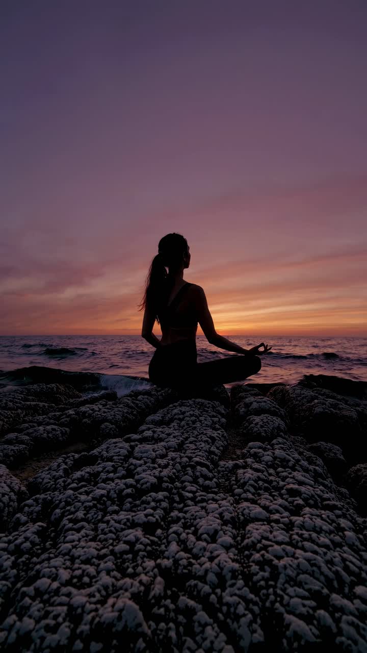 Silhouette of a person meditating on rocky shore at sunset, captured from a low angle