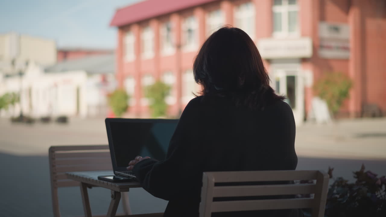Lady seated on wooden chair in open square working on laptop with smartphone on table against soft-focus blurred background of red building
