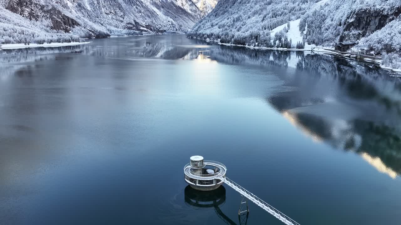 눈 덮인 알프스 산맥과 호수 위의 원형 급수탑이 보이는 클뢴탈러 호수의 고요한 겨울 항공 뷰