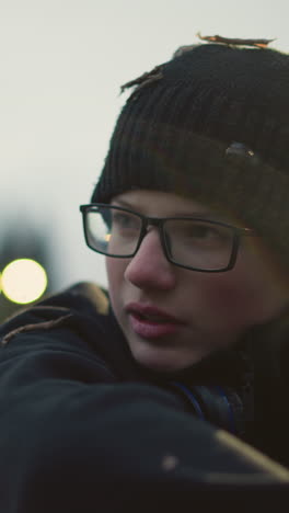 close-up of a young boy wearing glasses and a black beanie with grass particles on his head and headset on his neck, engaged in a discussion , with a blur background view