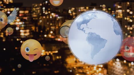 spinning globe and multiple face emoji icons floating against aerial view of cityscape at night