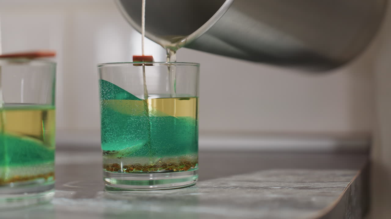 Elegant baker pours heated butter into third transparent cup placed on table beside two other filled containers with green decorative wax, wick held upright as golden mixture settles below surface
