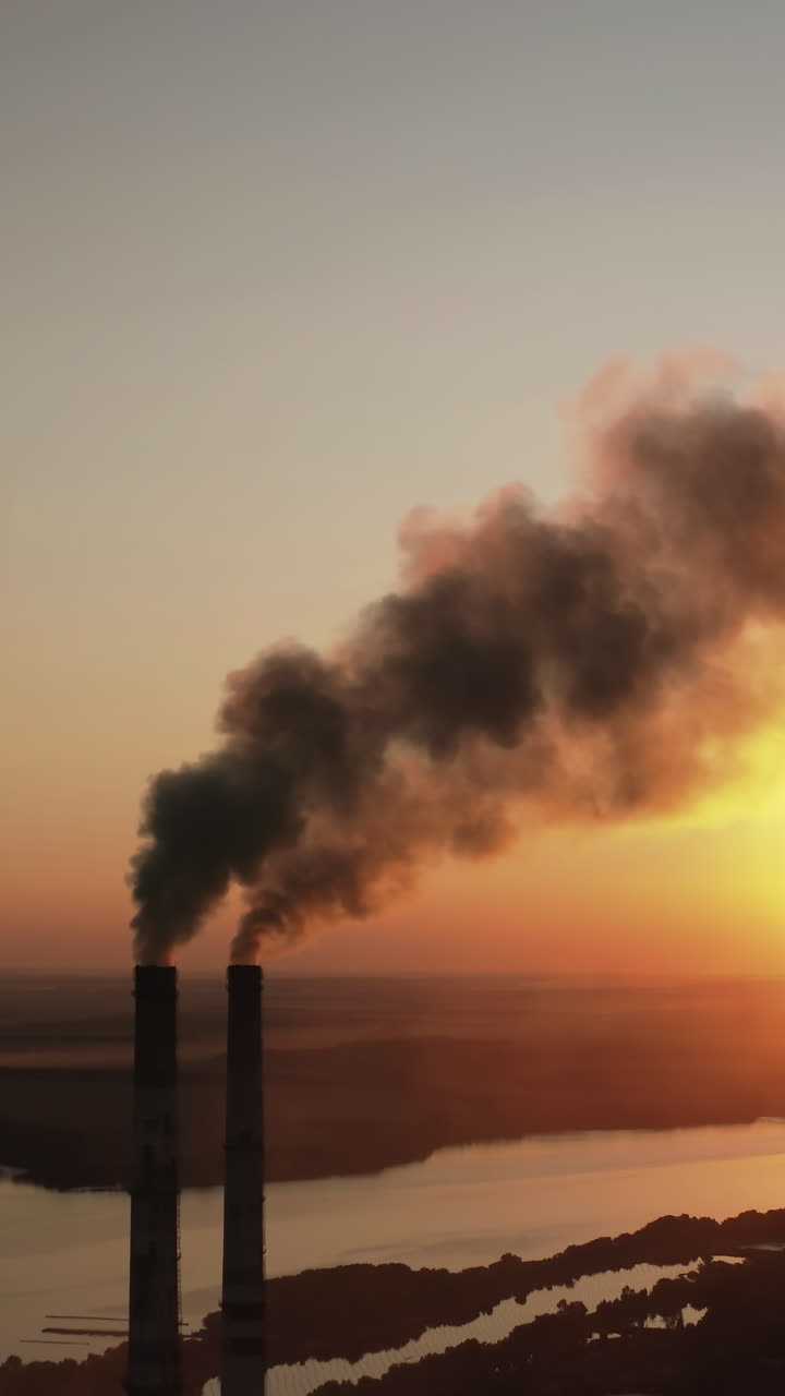 Factory pollutes environment at sunset. Toxic enterprise chimneys tubing against the evening sky background release black smoke. Smoking pipes of harmful manufacture in nature. Vertical video