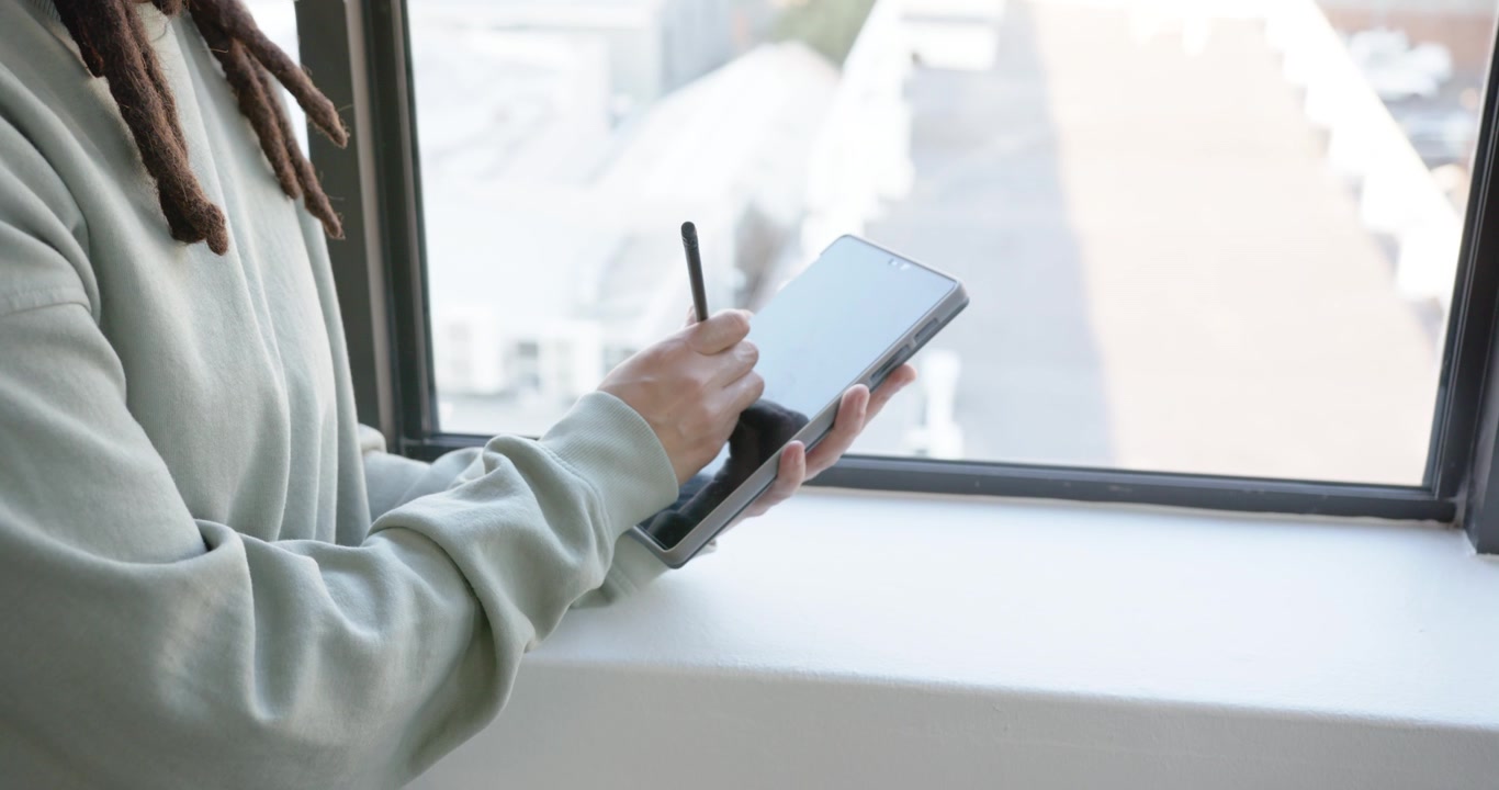 African American nonbinary in hoodie using stylus on tablet drawing at window, tilting, copy space