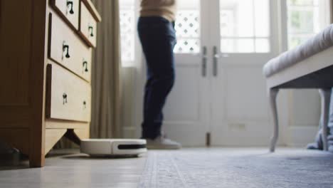 Midsection-of-caucasian-man-looking-through-window-with-robot-vacuum-cleaner-on-floor-in-bedroom