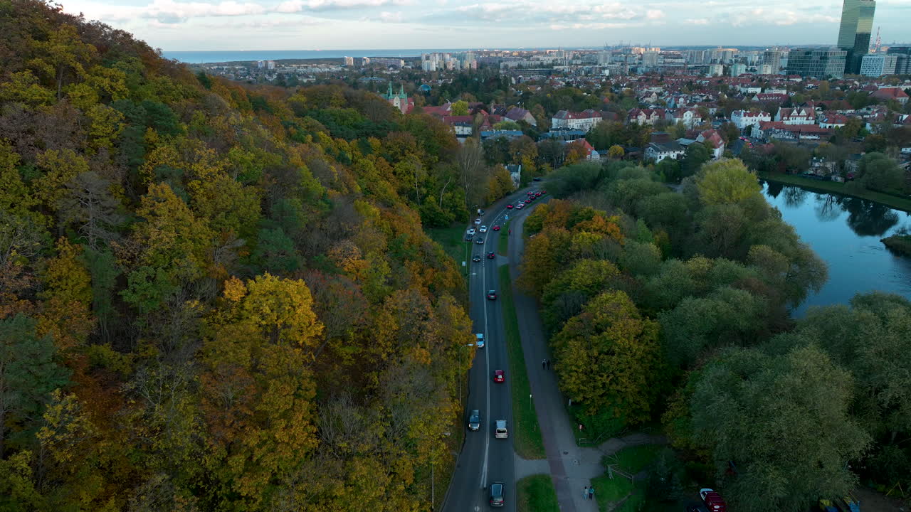High-angle drone view of a winding road through an autumn forest leading into a city with mixed historic and modern buildings - Gdansk, Oliwa