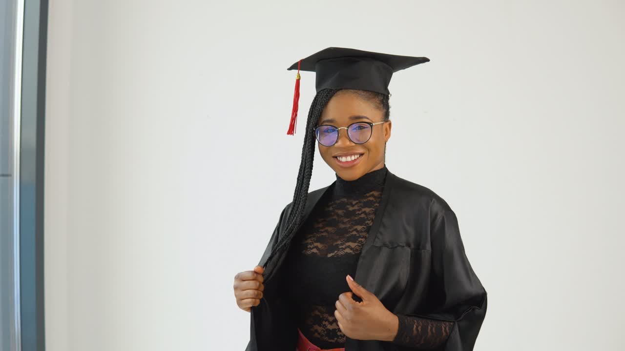 feliz sexy elegante mujer afroamericana estudiante en uniforme de posgrado de pie y coquetear en un fondo blanco