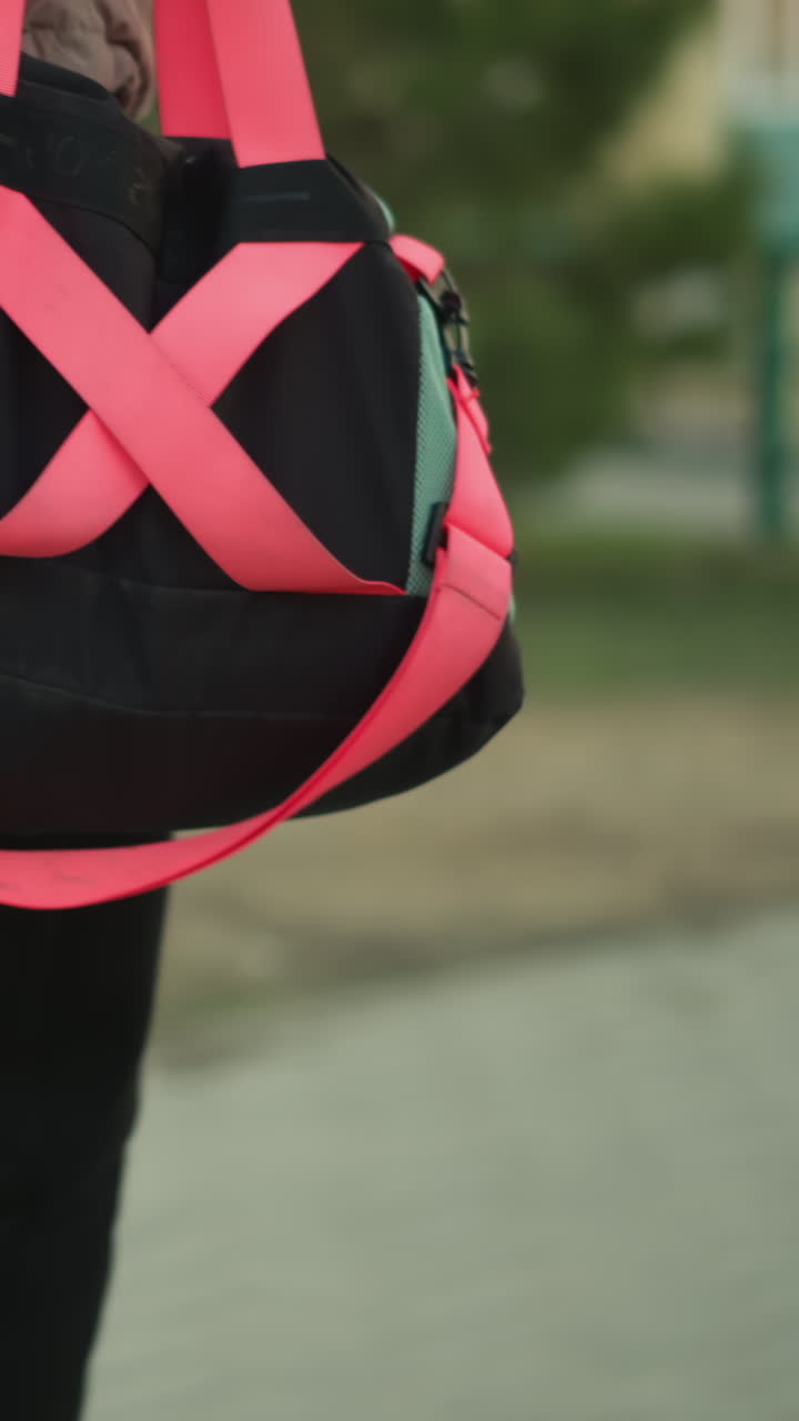 un primer plano de una chica vestida con una chaqueta de melocotón y pantalones negros, caminando por un parque mientras lleva una bolsa de gimnasia con correas rosas brillantes y un par de patines azules