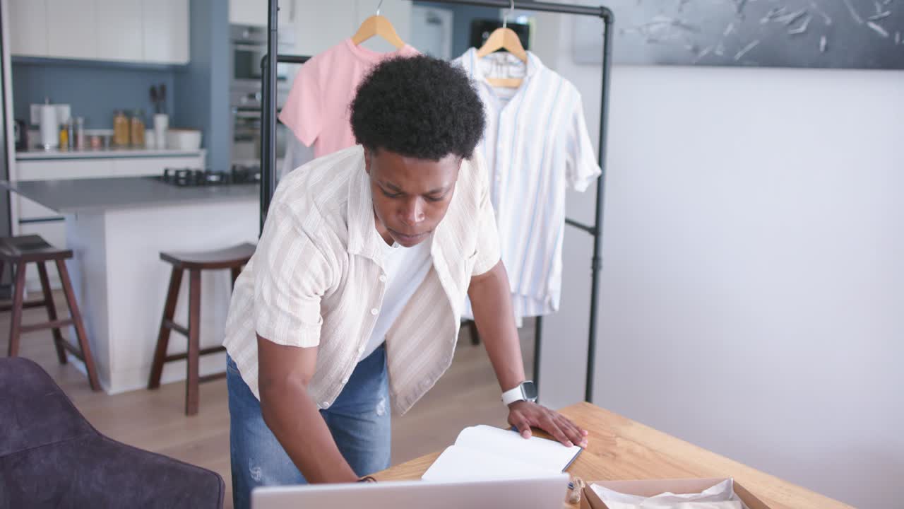 Apparel owner referencing laptop, flipping pages, writing, checking clothing rack to process orders