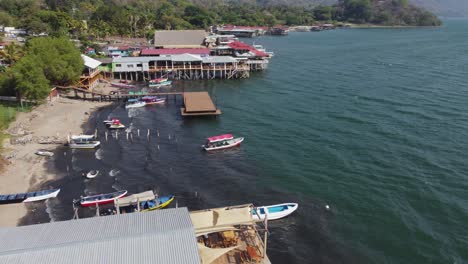 Flyover-docks,-buildings-on-crescent-beach-shore-of-Coatepeque-lake