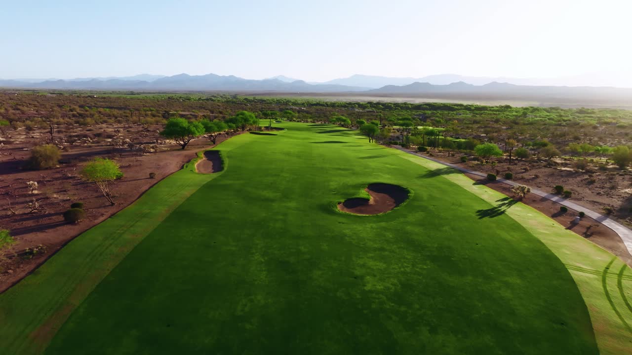 Push-in drone shot of a desert golf fairway with visible cart tracks, bunkers, and mountain views at sunrise