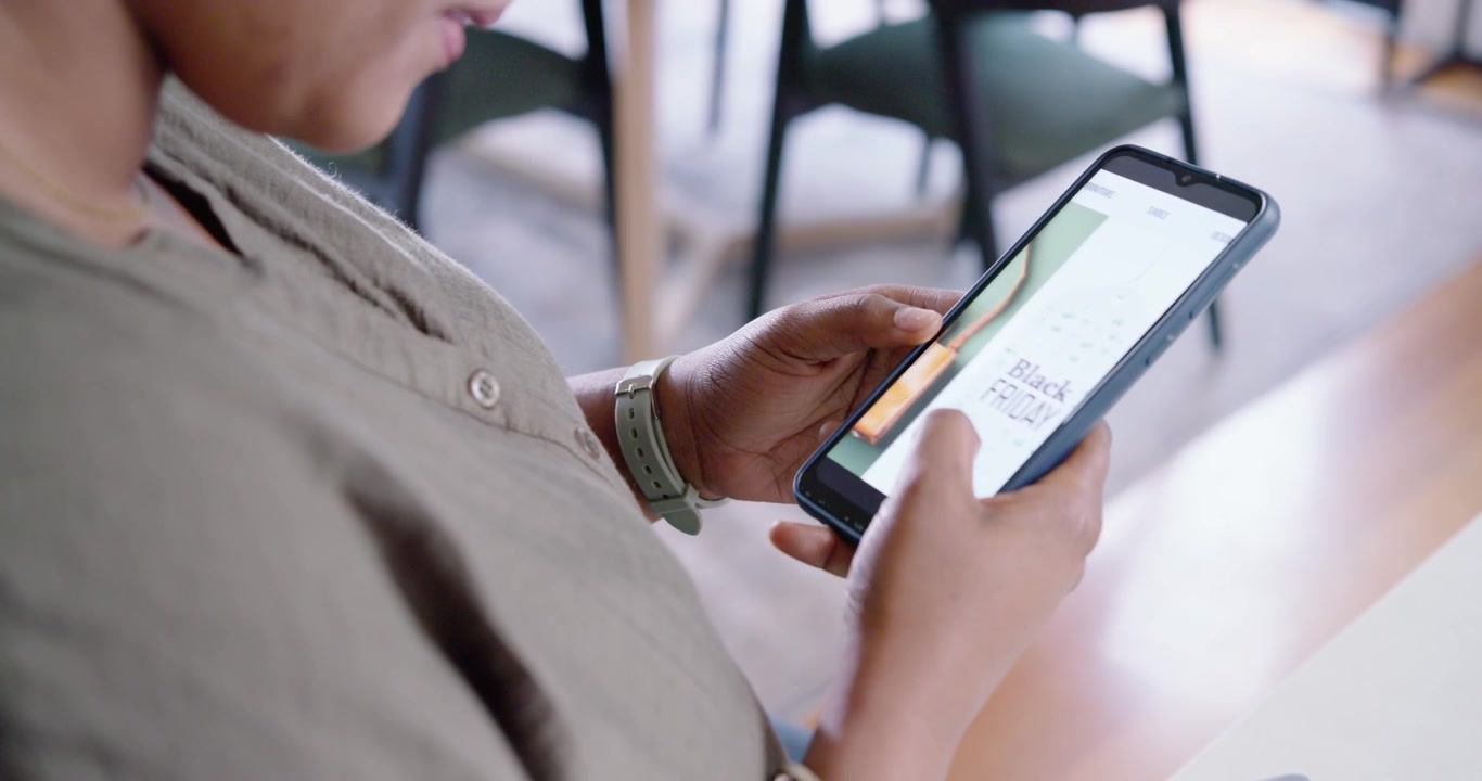 Woman wearing khaki shirt sitting in cafe holding card, scrolling phone, weighing Black Friday deal