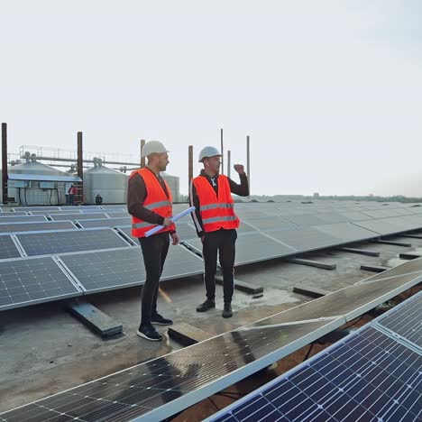 engineers talk on the solar farm background. rows of solar panels and two workers in orange uniform and hard hat. installation of innovative sunny batteries.