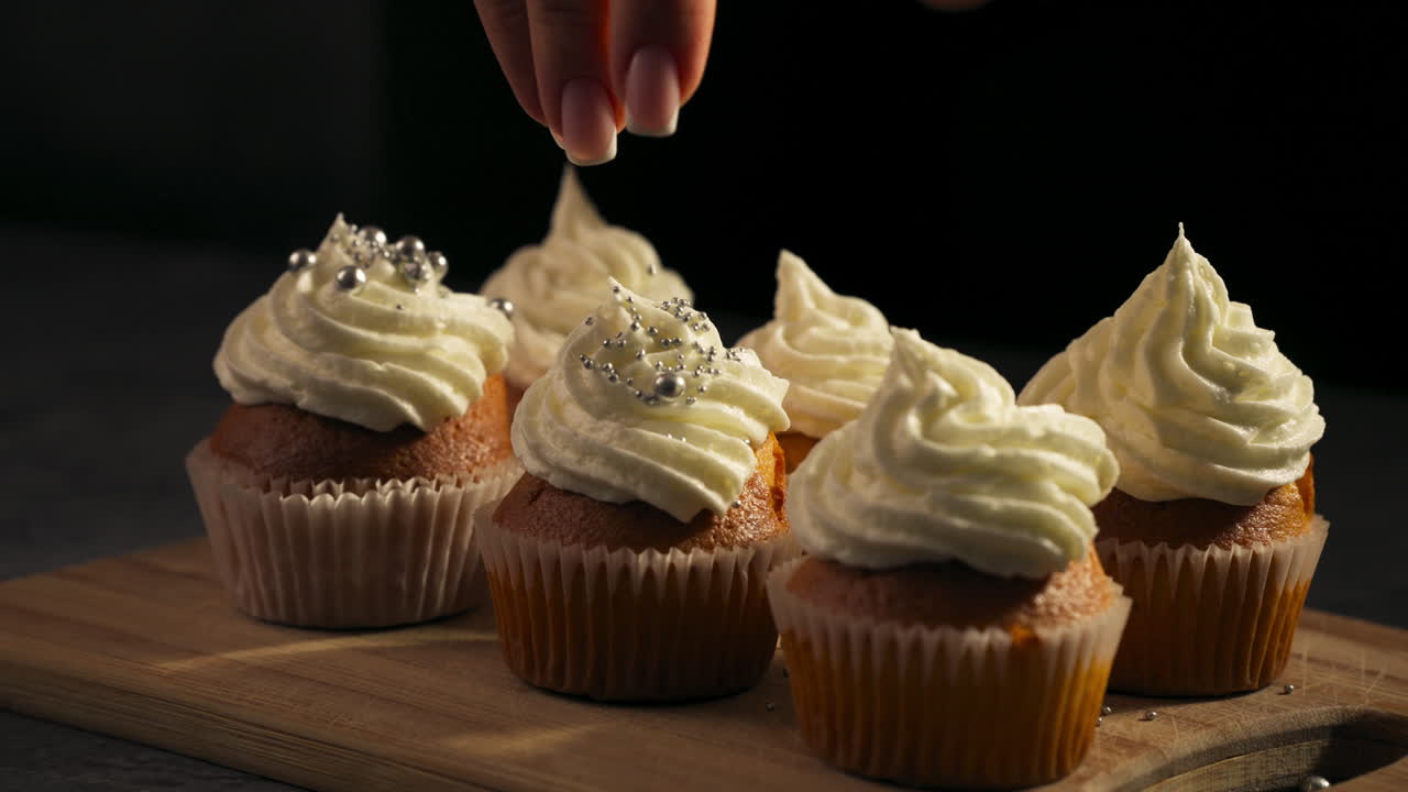 Perfectly frosted cupcakes neatly arranged on a wooden tray for presentation as hand drizzles sprinkles and shiny candy pearls