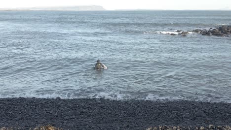 Valiente-Surfista-Se-Enfrenta-Al-Agua-Helada-En-Traje-De-Neopreno-Completo-En-Islandia,-Antena