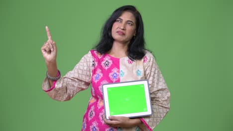 mature beautiful indian woman showing digital tablet and pointing finger up while thinking