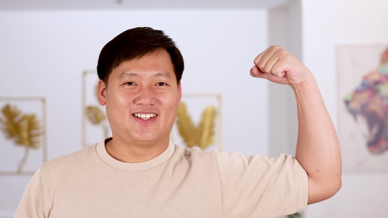 Asian man in beige shirt flexes arm and smiles confidently in a well-lit, contemporary indoor setting with soft, even lighting and steady camera