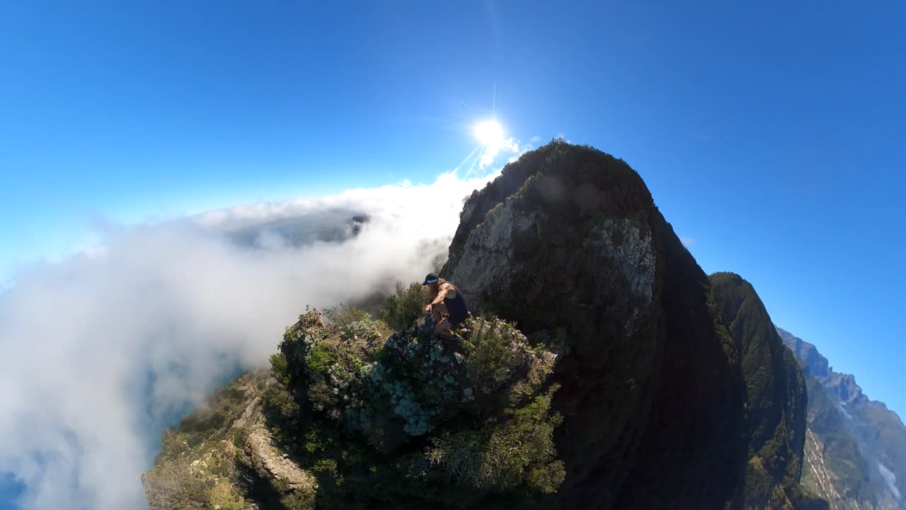 A young and strong solo hiker is walking along the steep edge of Espigao Amorelo in Madeira with thin clouds around him as he takes a dangerous step up to a rock on the cliffside to enjoy the view.