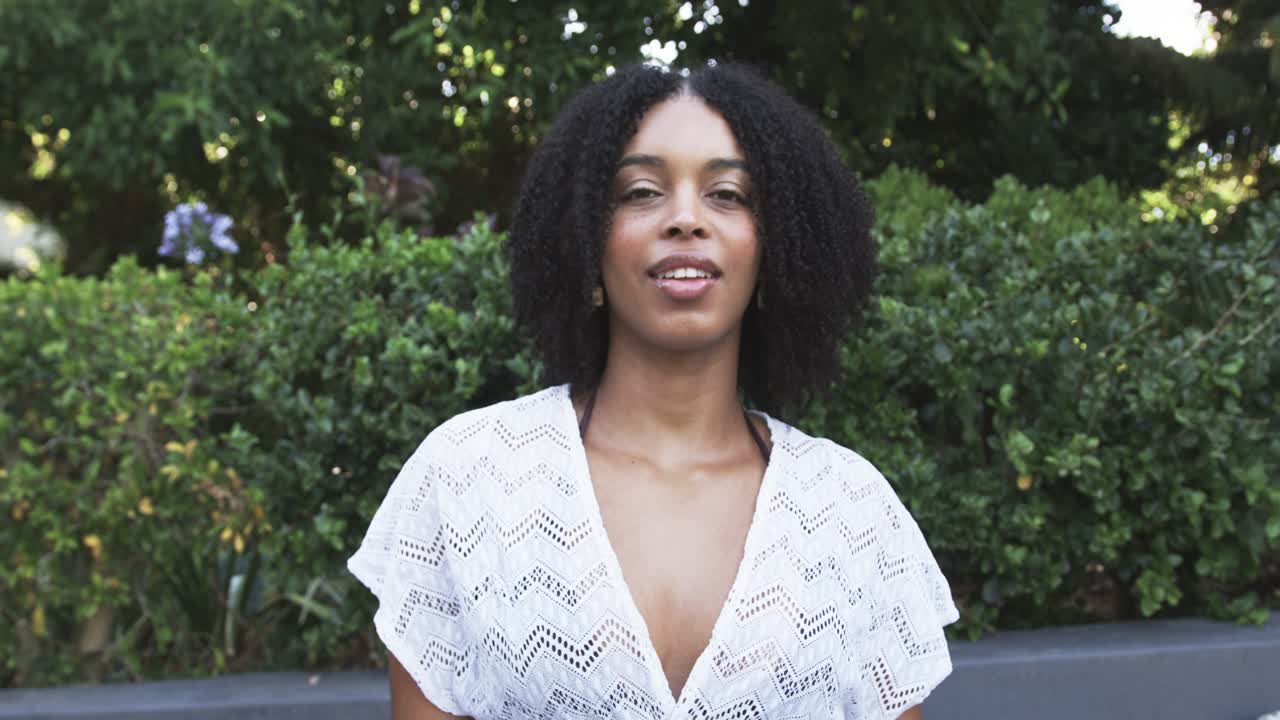 Afro-Amerikaanse vrouw glimlacht, raakt haar halslijn aan, past een witte kanten top aan, kijkt naar de camera bij een heg.