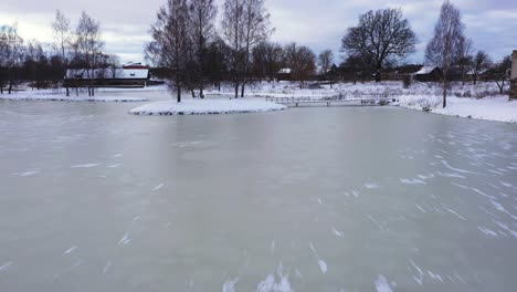 Frozen-Lake-in-Park-of-small-town-with-snowy-landscape,-aerial-low-altitude-view