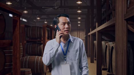 asian male professional worker standing in cellar of winery, he pick the phone up and started to speaking with it