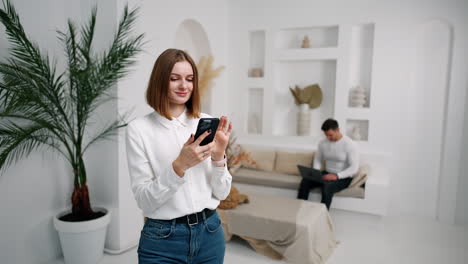 brunette caucasian girl in white shirt focused on her smartphone. lady raises her gadget to take selfie indoors. man sits at backdrop working on laptop.