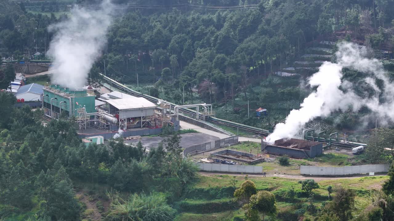 Aerial view of a geothermal power plant surrounded by lush green hills, showing steam rising from turbines and pipelines generating renewable energy