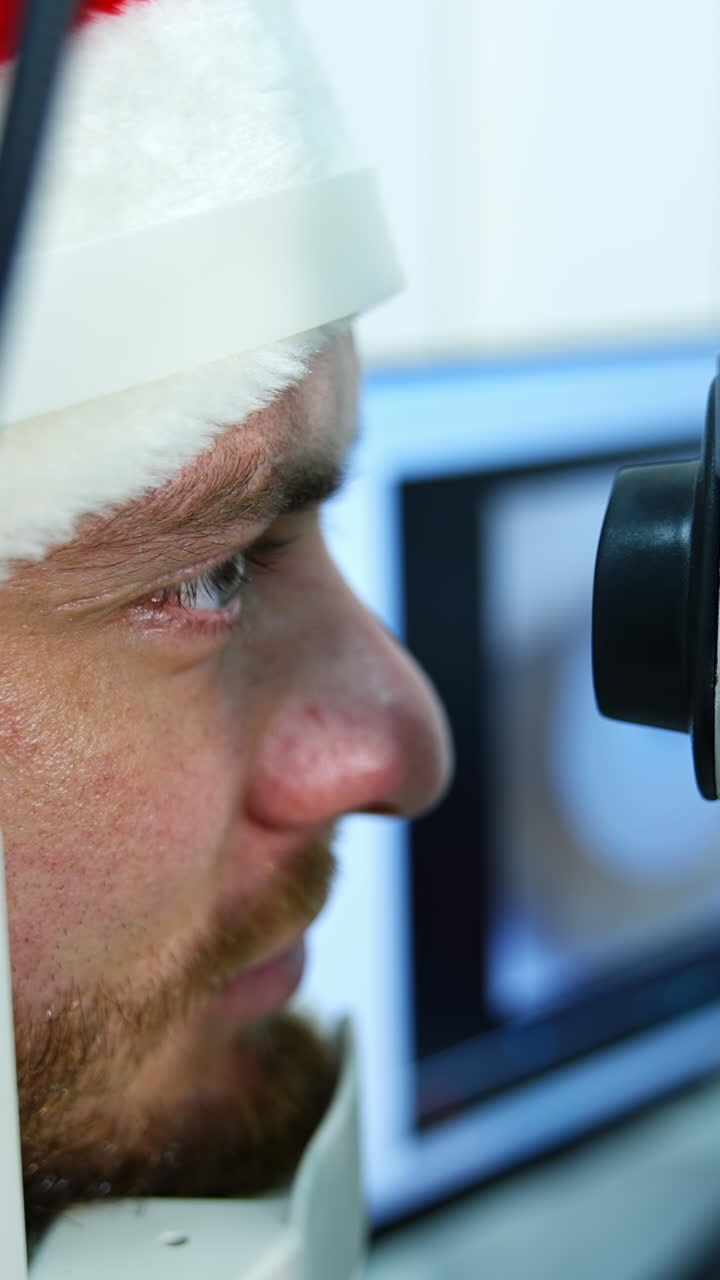 Patient treating eyes in clinic. Profile view of a man's face in Christmas cap looking into special device in ophthalmology center. Close-up. Vertical video