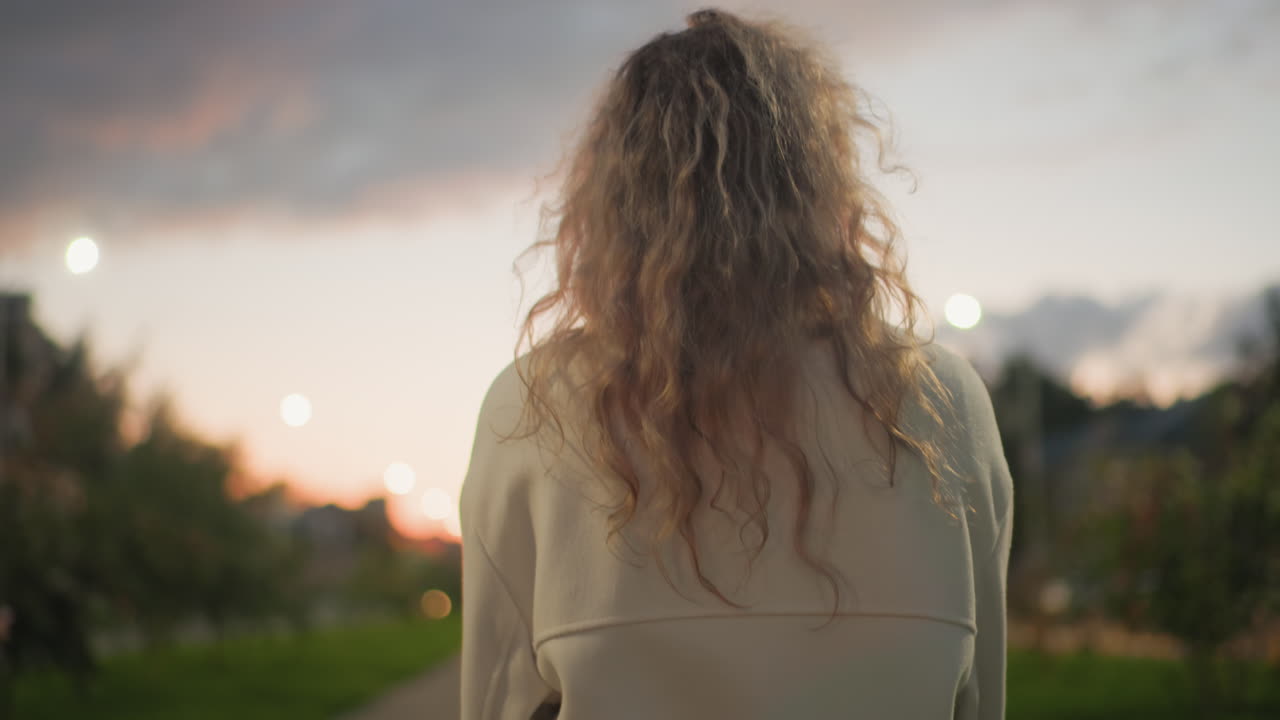 Back view of woman in white coat rubbing her hands together during peaceful evening stroll with sunset glow and soft bokeh lights creating dreamy atmosphere over blurred park and residential area