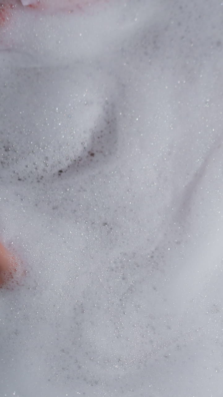 mujer cubierta de espuma esponjosa yace en la bañera en casa. dama disfruta de la relajación y el procedimiento de spa en el baño. mujer mima el cuerpo en agua caliente