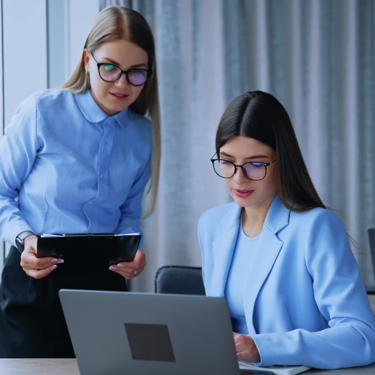 Female office colleagues having conversation while looking at the screen of laptop. Busy young ladies in glasses discussing work issues