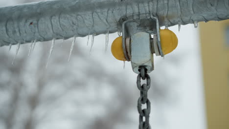 close-up of metallic swing chain joint with yellow stopper coated in ice and icicles hanging along metal bar, highlighting frozen textures and wintry detail against a blurred snowy background