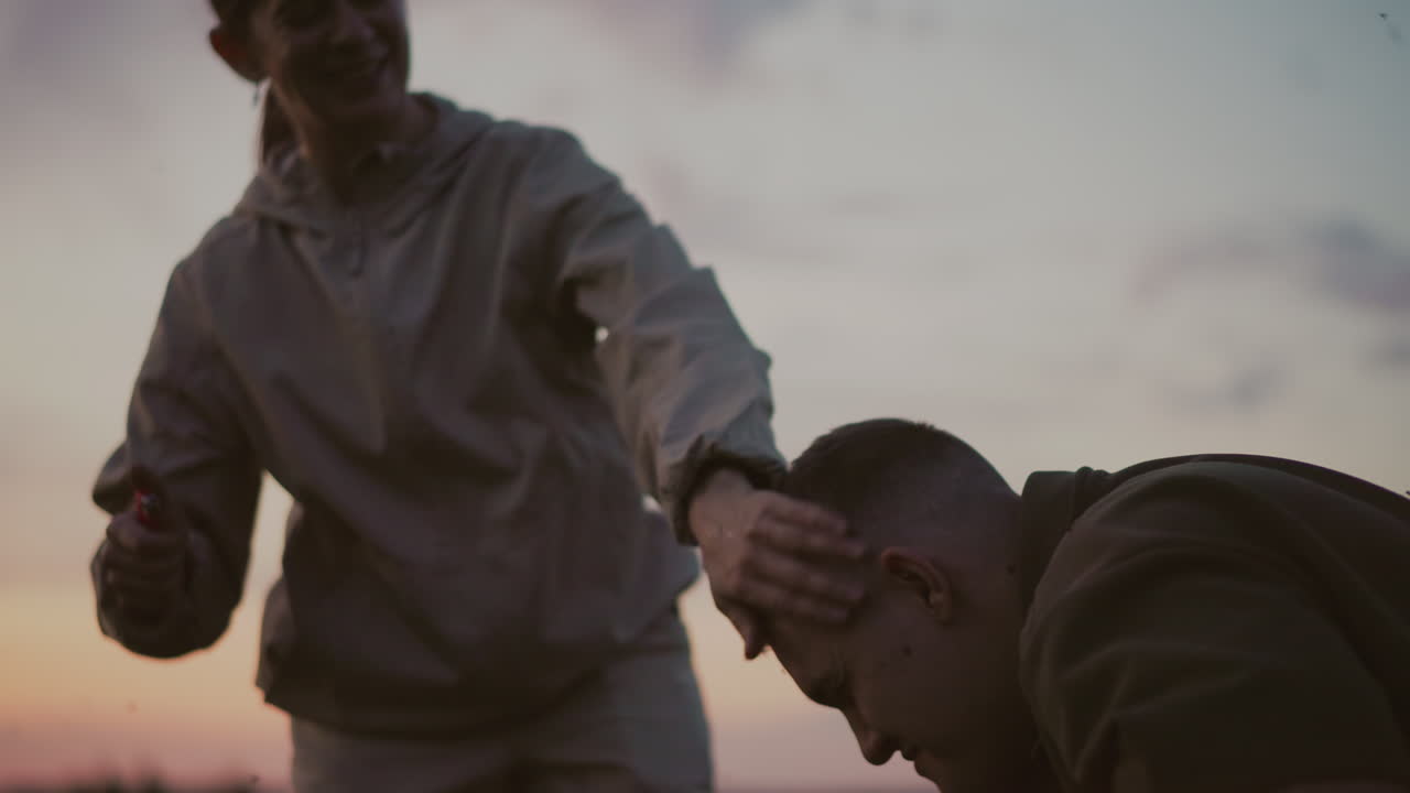 close up of woman holding lighter against man hair tip while water pours from bottle to quench flame in sunset field setting capturing tense instructive moment with protective intervention