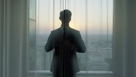 silhouette of a man in a suit standing by a window with city skyline at sunset