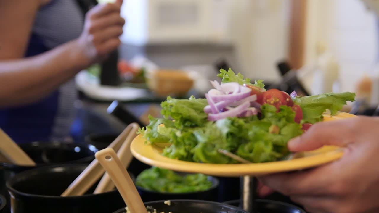 A man serves himself salad from the cafeteria salad bar, placing it on a plate in slow motion. Other people are in the background.