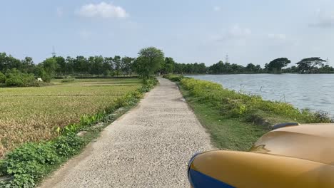 portion of a yellow bengal taxi standing near a village road with pond and agricultural fields on both side