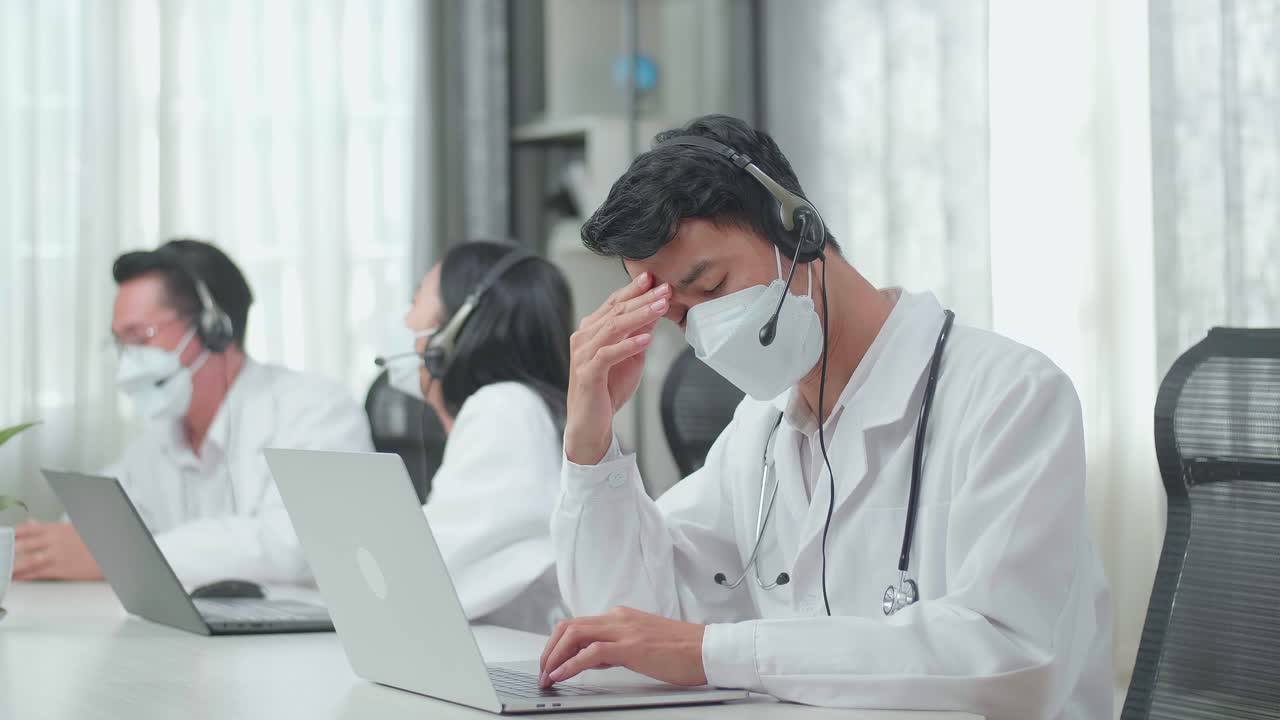 A Man Of Three Asian Doctors With Stethoscopes In Headsets And Masks Working As Call Center Agent Is Tired Because His Colleagues Are Talking During Working At The Office