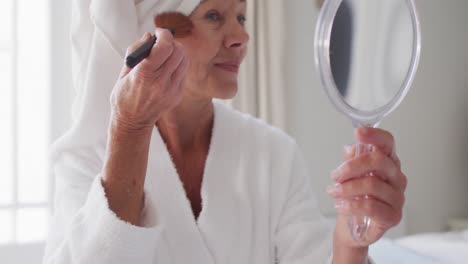 Happy-senior-caucasian-woman-sitting-on-bed-in-bedroom,-putting-on-make-up