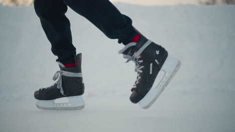 side view skater in black ice skates gliding smoothly across frozen rink snowbank background visible with soft winter light creating contrast between skates and icy surface