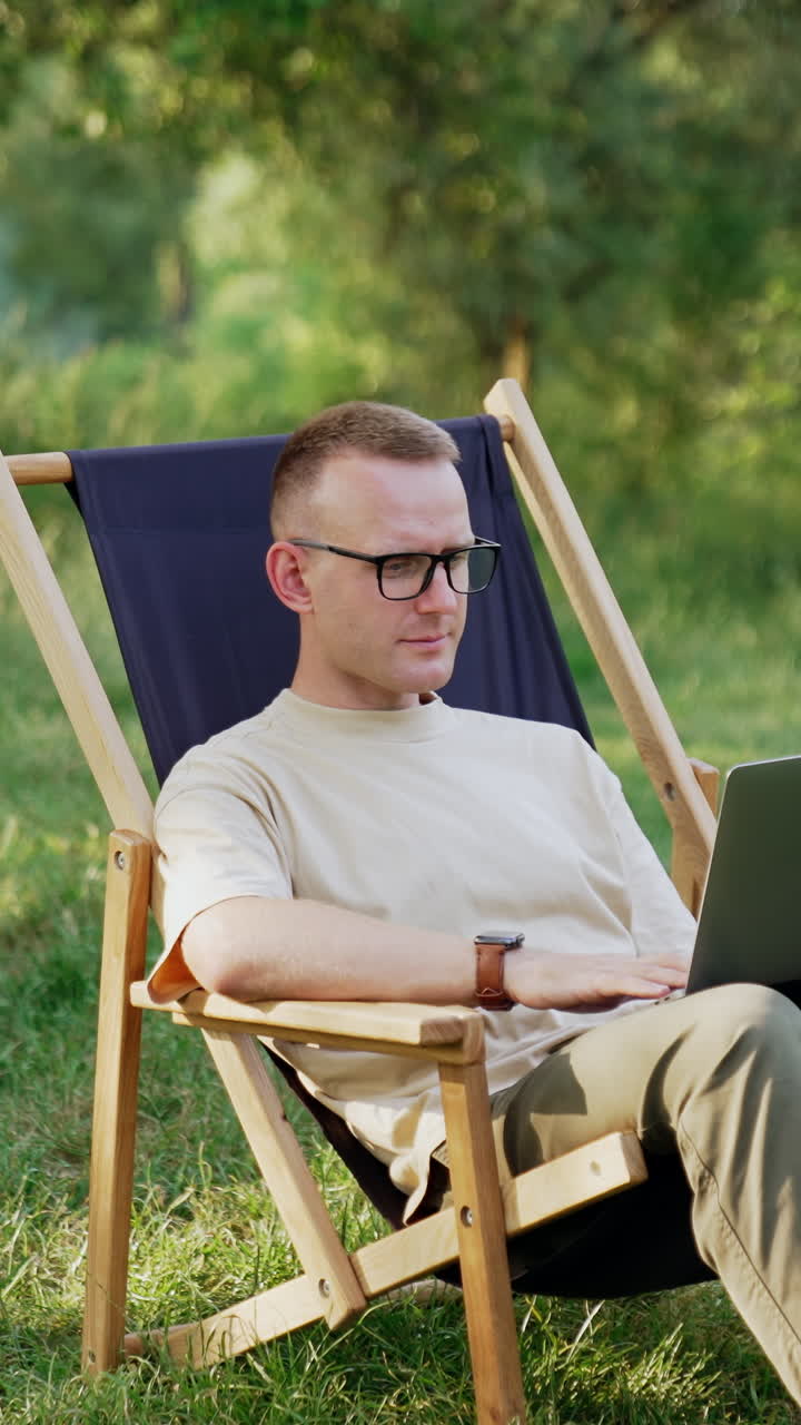 Caucasian freelancer starting work on laptop. Man opens laptop sitting in chair on the meadow. Car at backdrop. Vertical video
