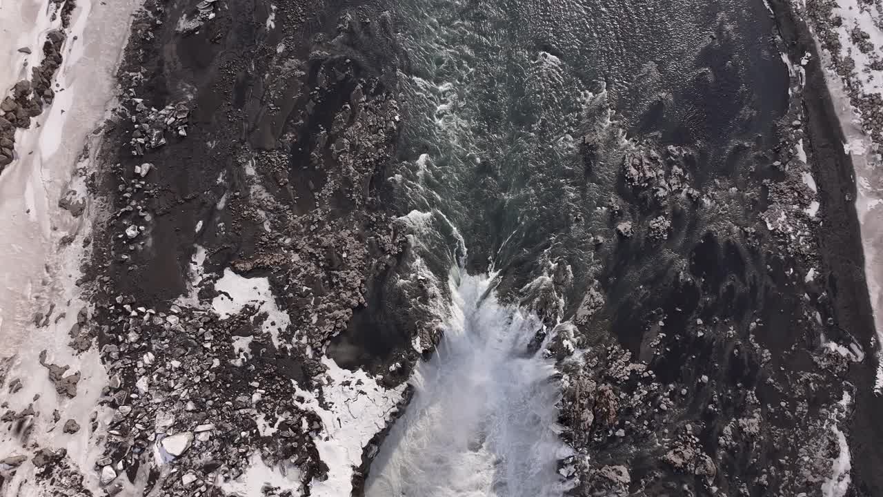 Top-down aerial view of a dramatic waterfall plunging into dark basalt terrain near Selfoss, Iceland, showcasing raw energy and contrast in a rugged winter landscape.