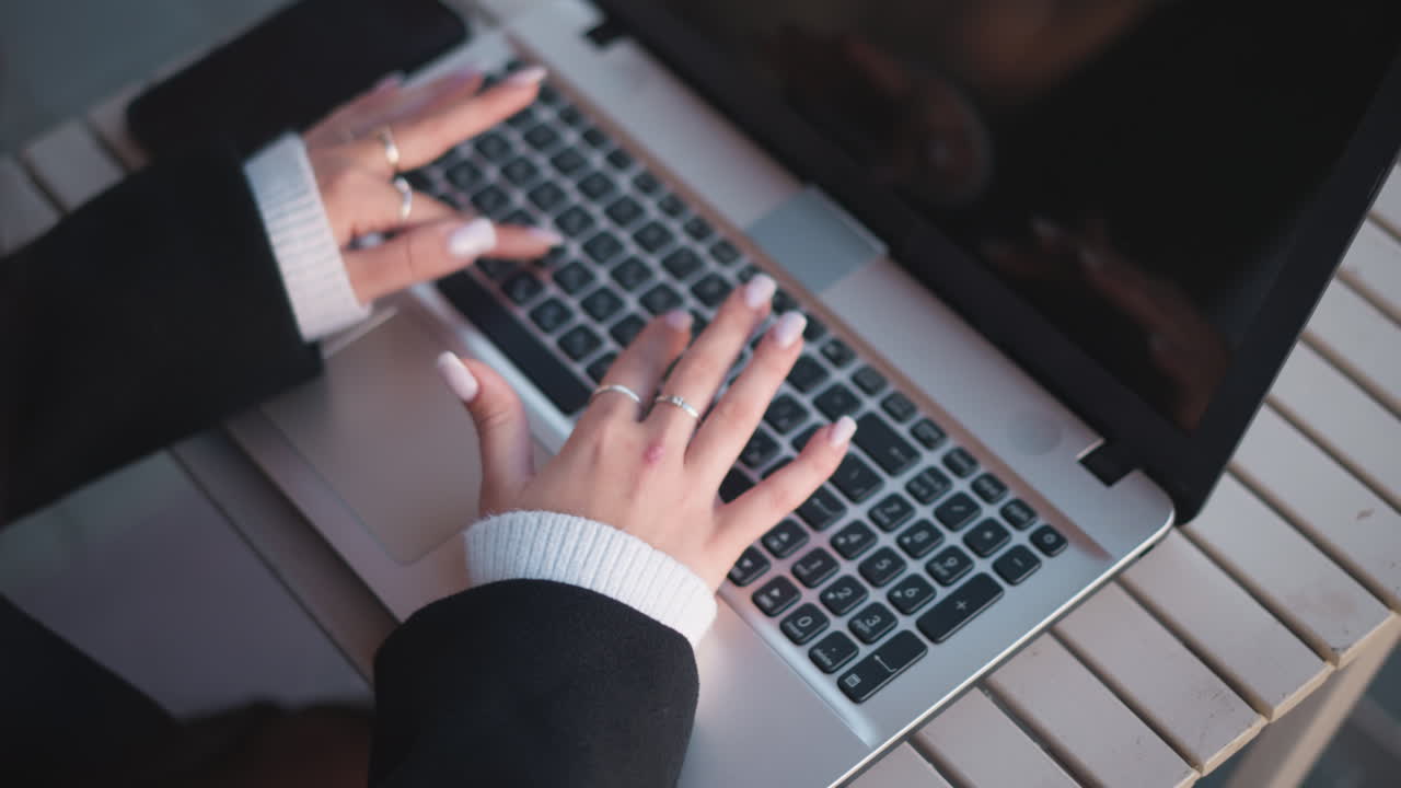 primer plano de manos escribiendo en el teclado de la computadora portátil, adornado con anillos de plata y clavos pulidos, cómodas mangas de suéter visibles, con el teléfono inteligente colocado cerca de la laptop en una mesa de madera