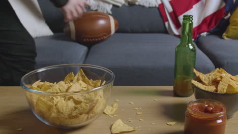 Persona-En-Casa-Viendo-Un-Partido-De-Fútbol-Americano-En-La-Televisión-Con-Bebidas,-Bocadillos-Y-Pelota-2