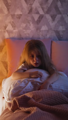 frustrated little girl with straight hair holds soft cushion and stares into vacancy sitting on bed near illuminated play tent in bedroom slow motion